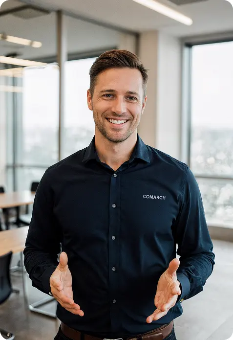 Uśmiechnięty mężczyzna w ciemnej koszuli z logo Comarch stoi w biurze.